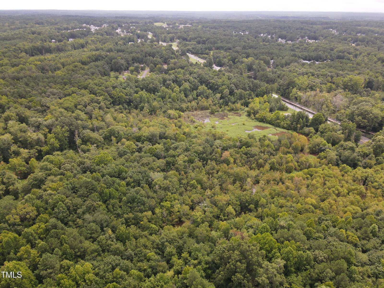 Aerial view of a vast forested area with dense green trees. A road cuts through the landscape in the upper right section, surrounded by a mix of open and wooded areas under a cloudy sky.
