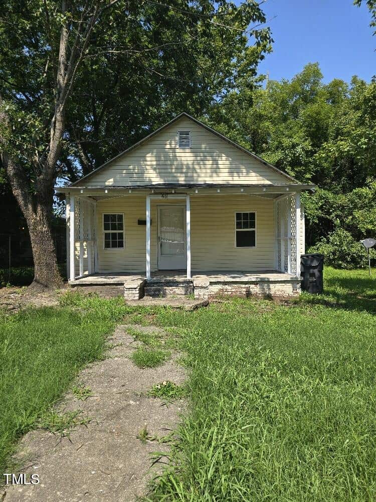 A small yellow house with a white front door and porch is surrounded by greenery at 611 Rowland Street. The house has a slanted roof, with a tree on the left side. A narrow stone pathway leads up to the house, set against lush grass and bushes in Henderson. A black trash bin is to the right.