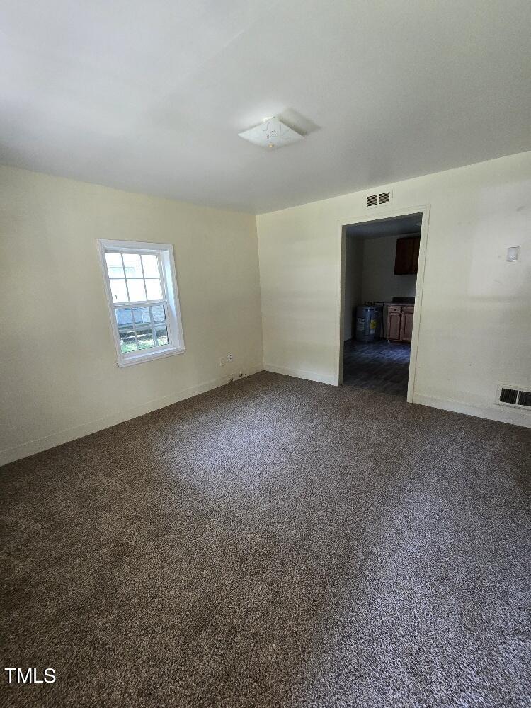 A small, empty room with beige carpet and light-colored walls. A white-framed window allows natural light in from the left wall. A narrow doorway leads to a kitchen area with visible cabinets and a water heater. A ceiling light fixture is centered on the ceiling.