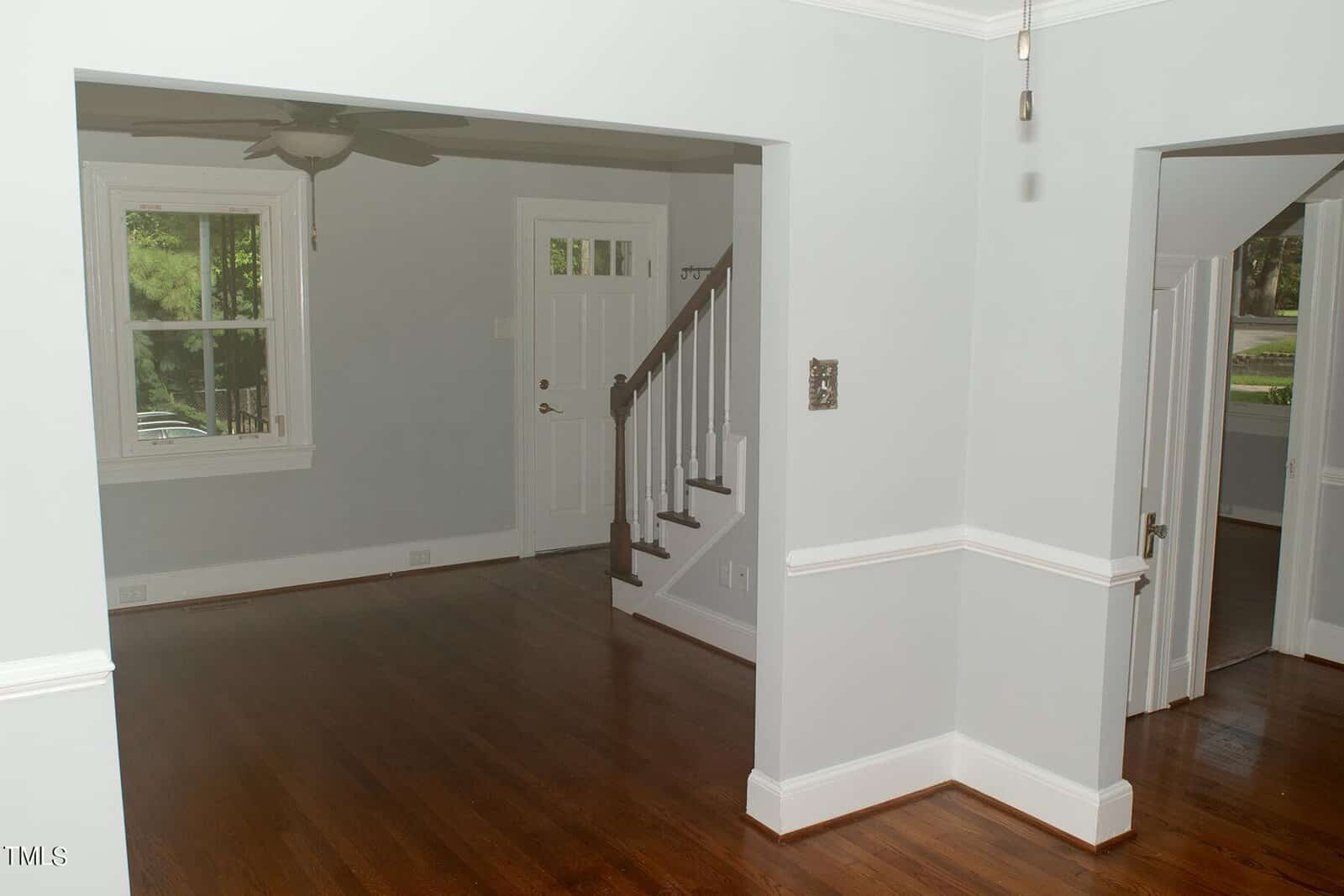 A room with hardwood floors, light gray walls, and white trim. There's a window on the left, a ceiling fan, and a view of a hallway leading to a staircase and a white door. Natural light comes through the window.