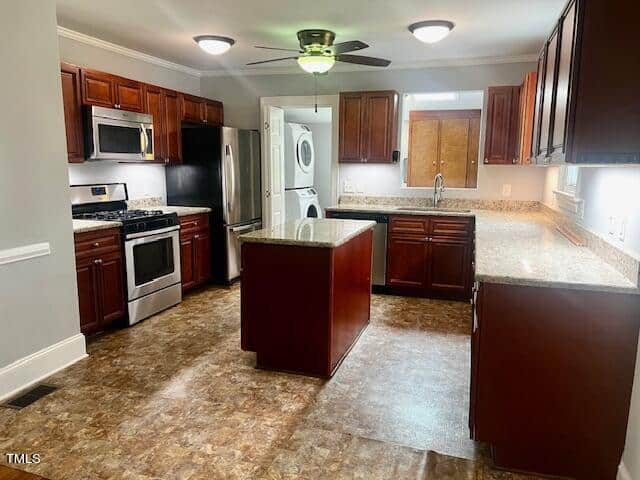 A modern kitchen with dark wood cabinets, stainless steel appliances, granite countertops, and a central island. A ceiling fan is mounted above, and a stacked washer and dryer are visible in the laundry nook. Floors are a mix of tile and laminate.