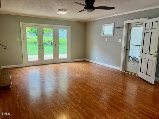A spacious, empty living room with wooden flooring and light gray walls. It features a ceiling fan, large glass doors leading to a grassy outdoor area, and a windowed door on the right side. Natural light fills the room through the doors and windows.