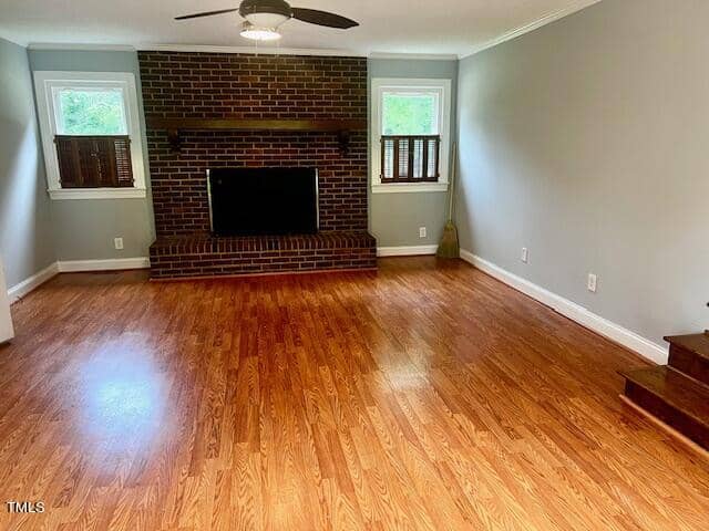 A cozy living room with polished hardwood floors, a brick fireplace centered between two windows with shutters, neutral gray walls, and a ceiling fan. A broom rests against the wall near a set of stairs.