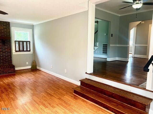 A spacious living area with light wood floors, neutral wall colors, and a brick fireplace. The room features a step-up to an adjoining space, a ceiling fan, and windows letting in natural light.