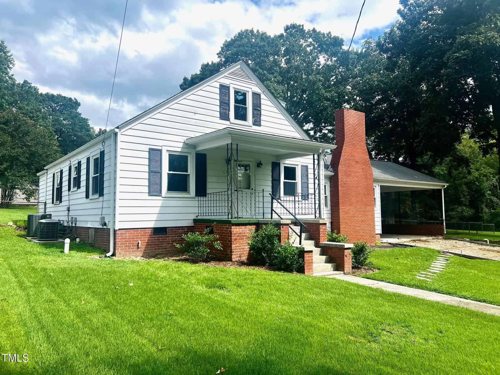 The charming house at 111 North Street, Oxford, is a white, one-story dwelling accented with a red brick chimney and stairs. It boasts black shutters and a quaint porch with white railings. A green lawn and partially visible driveway complete the scene, framed by trees under a cloudy sky.