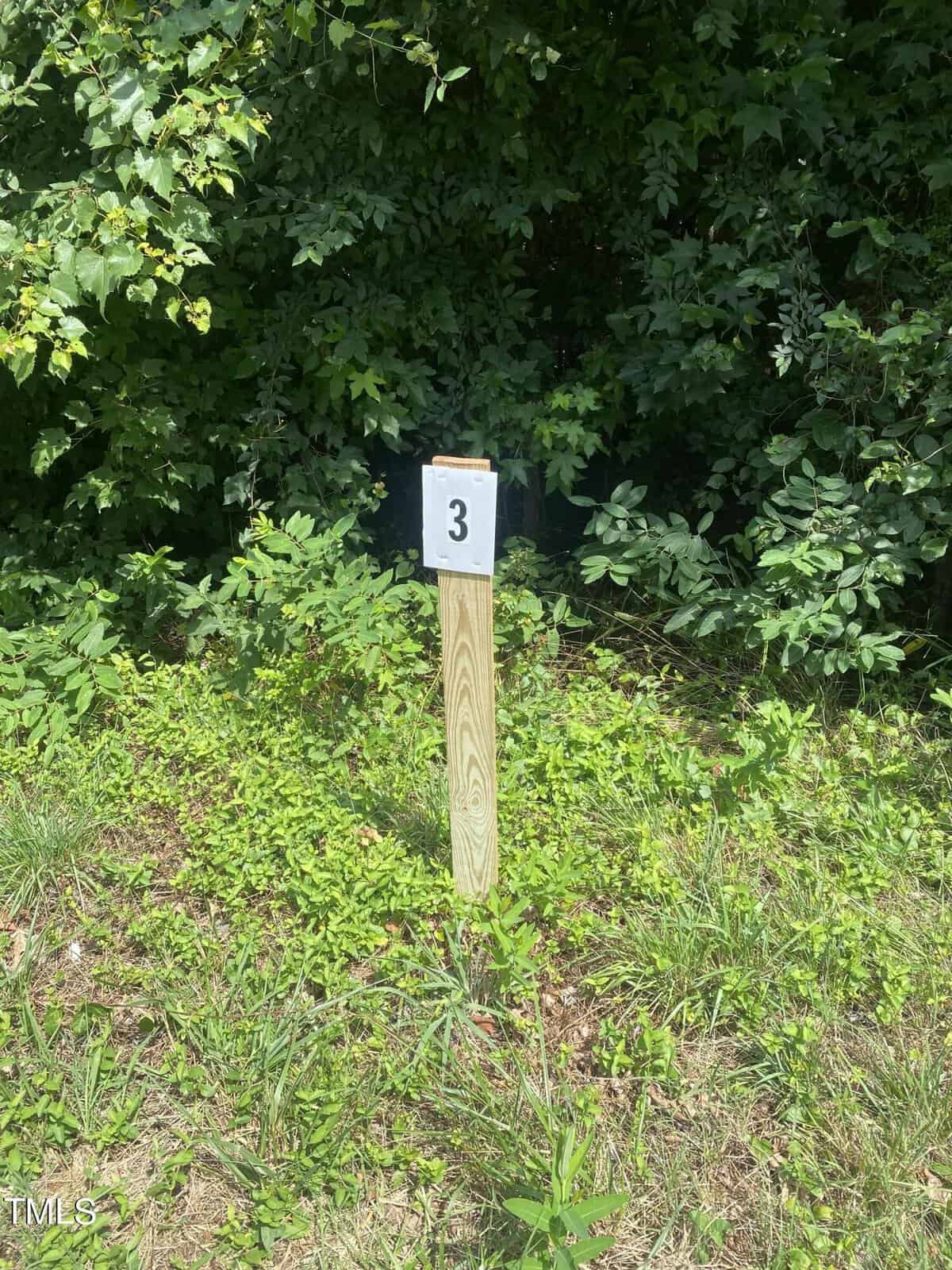 A wooden post stands surrounded by greenery, with a white sign displaying the number "3" attached to it. Dense foliage and grass fill the background.