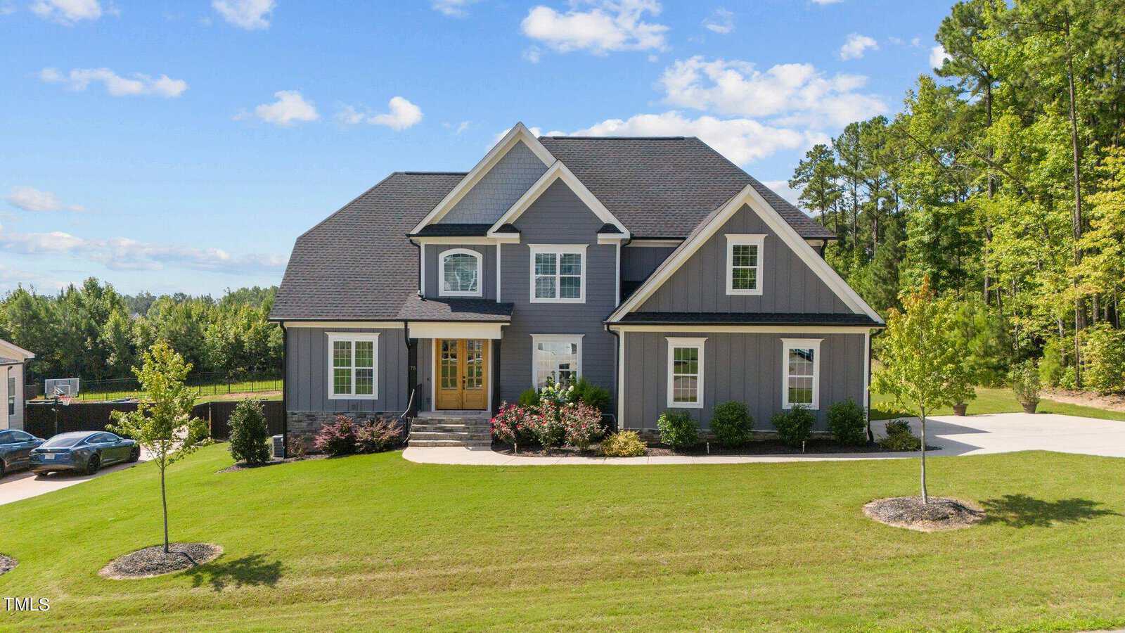 A modern two-story gray house with white trim sits on a neatly manicured lawn, surrounded by small trees and shrubs, under a bright blue sky with scattered clouds.