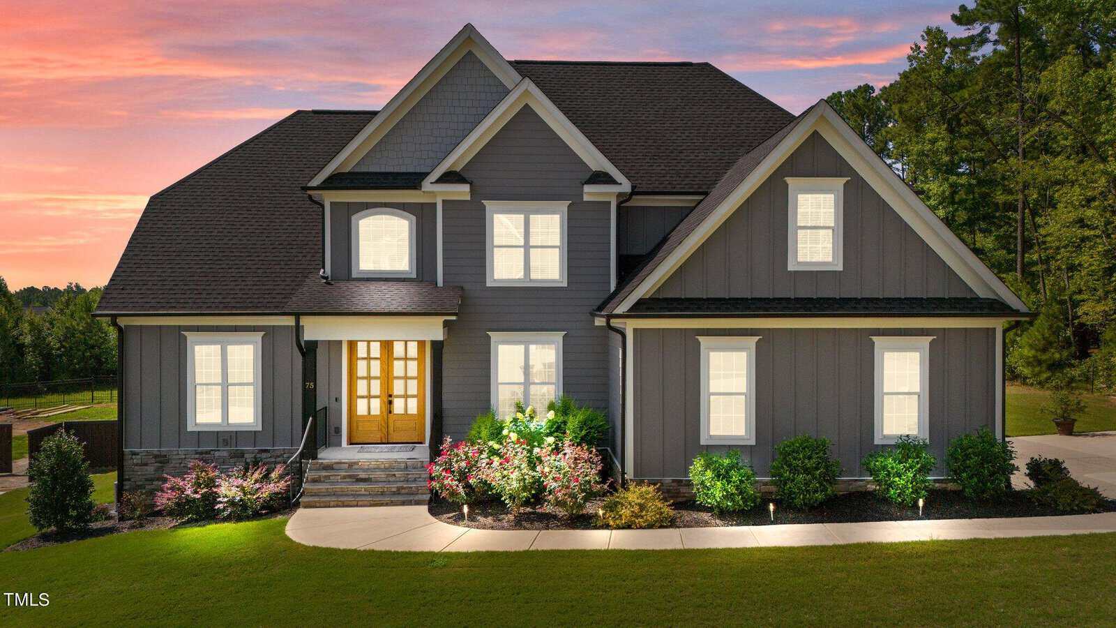 A modern two-story gray house with white trim, large windows lit from within, a wooden front door, and a landscaped front yard with shrubs and flowers, set against a sunset sky and surrounded by trees.