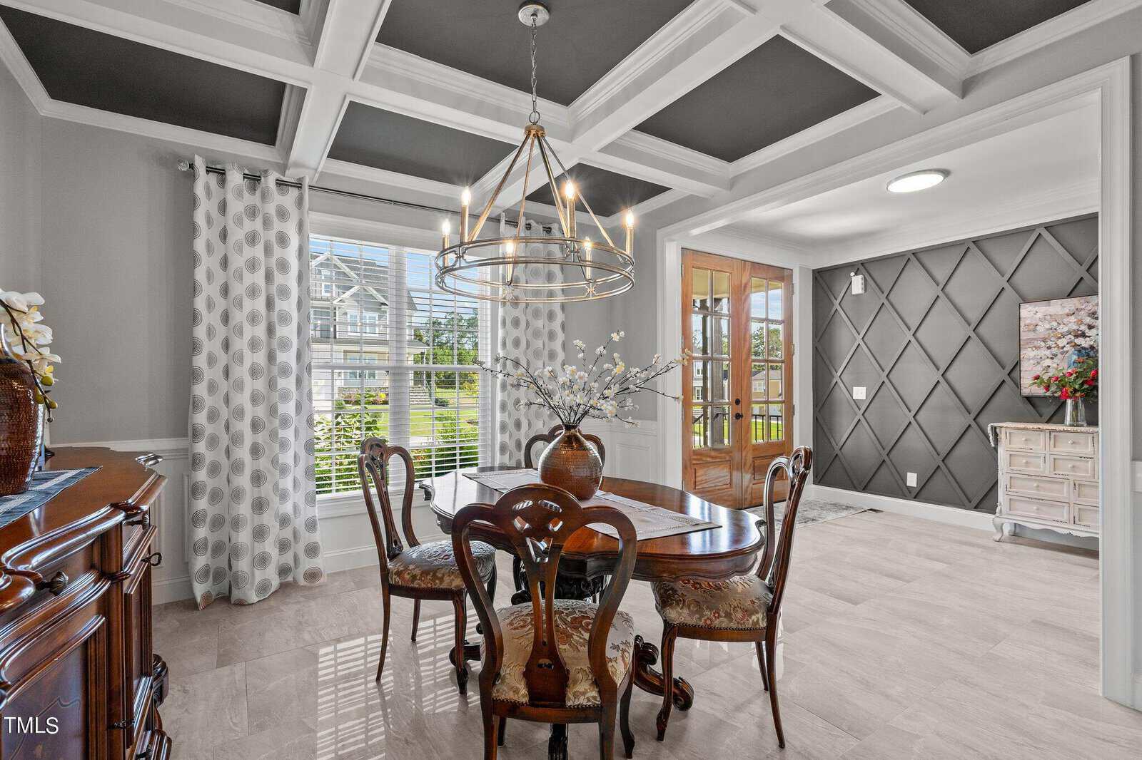Elegant dining room with a wooden table and four chairs, a modern chandelier, coffered ceiling, large window with patterned curtains, and an open entryway with decorative wall paneling.