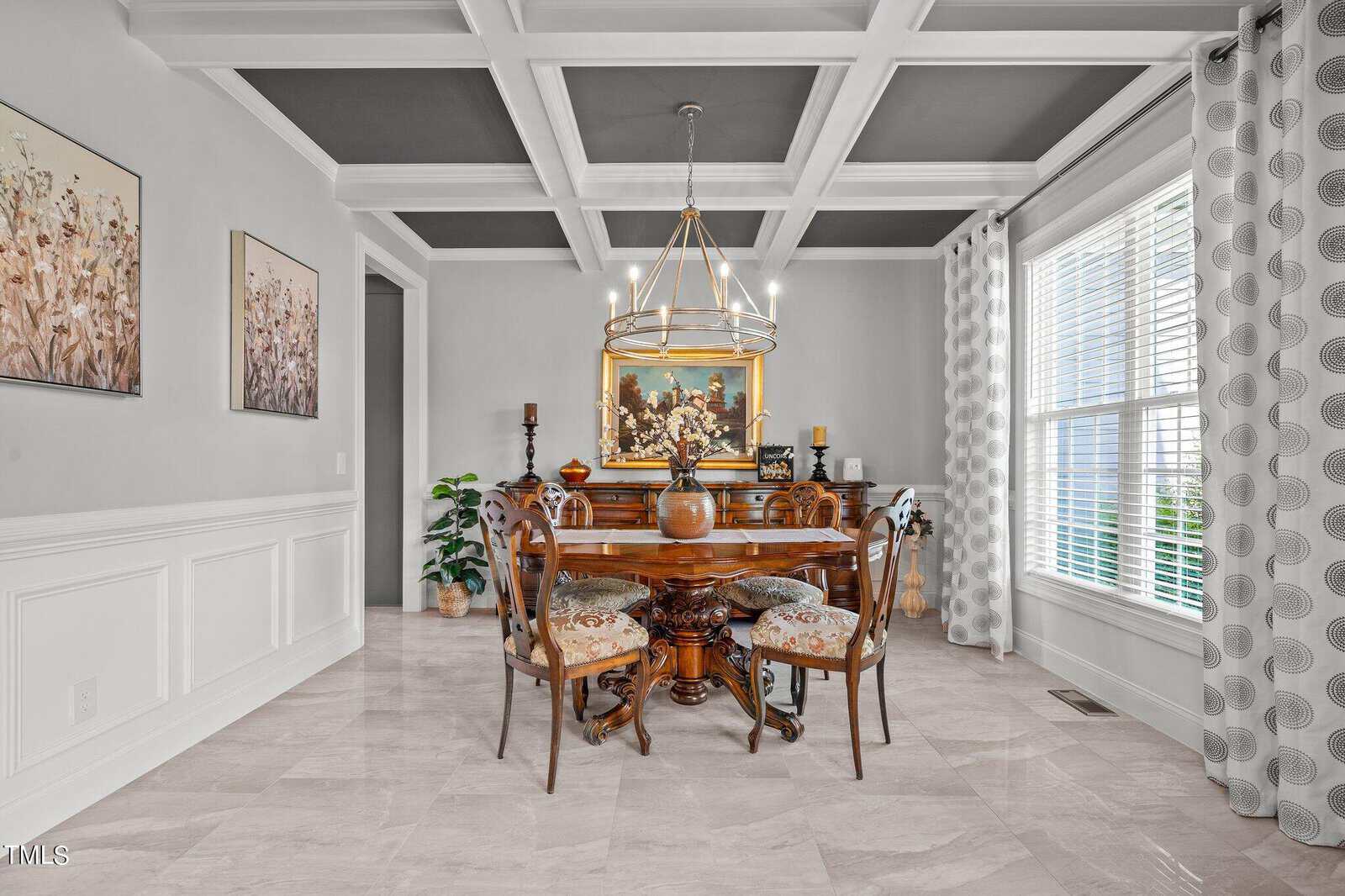 Elegant dining room with a coffered ceiling, a round wooden table with four ornate chairs, a chandelier, large window with patterned curtains, and artwork on light gray walls. A piano sits against the far wall.