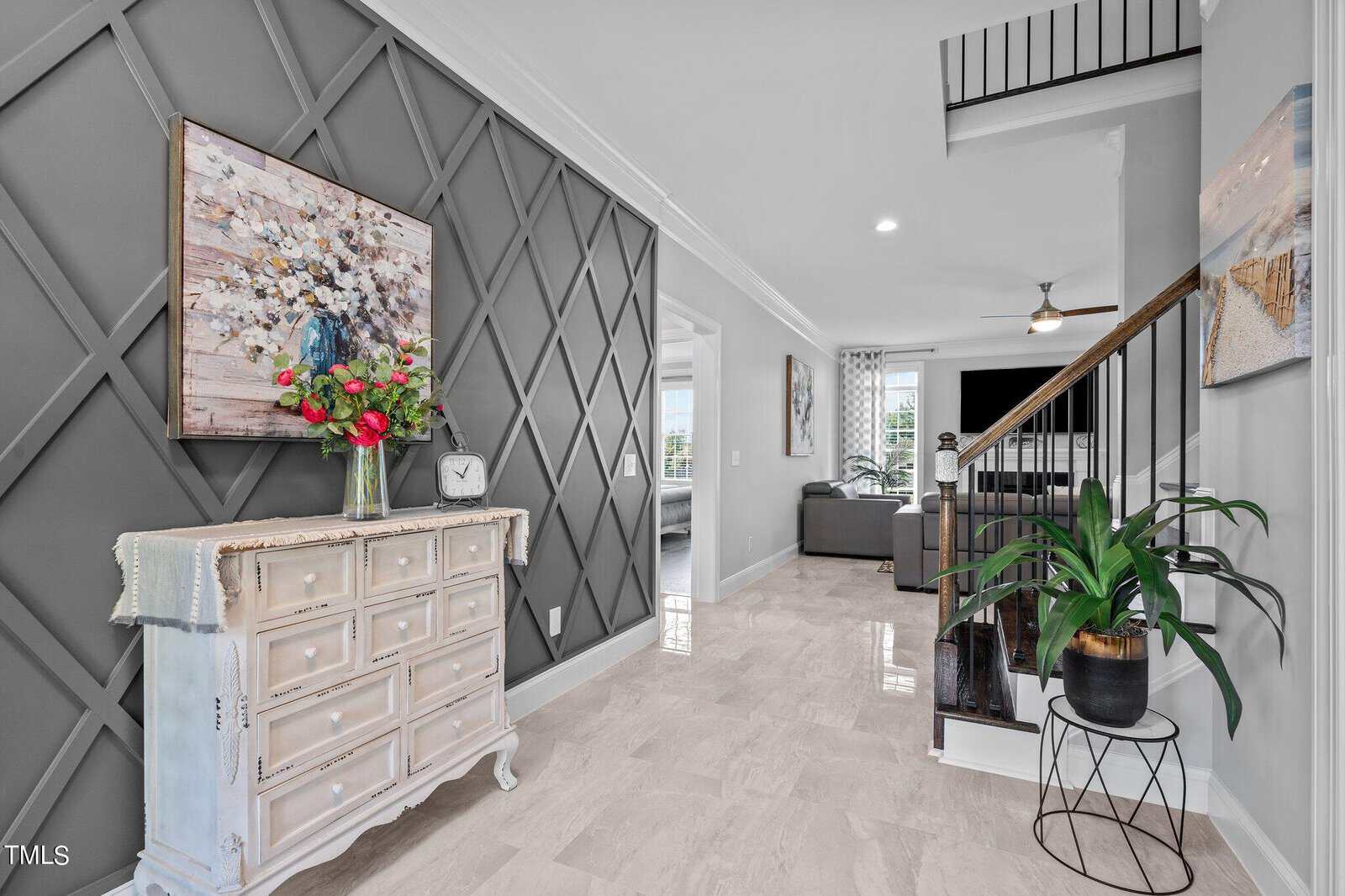 A bright, modern entryway with light marble floors, a gray accent wall featuring geometric patterns, a white vintage chest with flowers, stairway with black railing, wall art, and a potted plant on a metal stand.