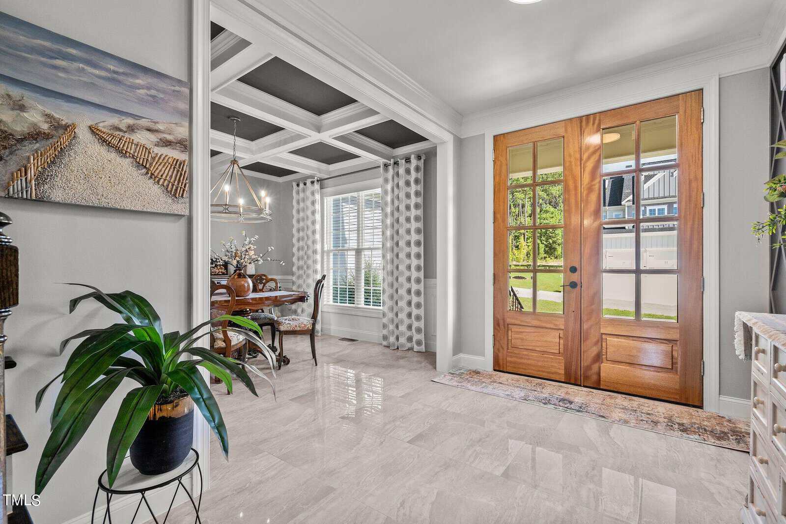 Bright entryway with wooden double doors, a small rug, and a potted plant on a stand. To the left, a dining area with a table, chairs, and a chandelier is visible through an open doorway. Large windows let in natural light.