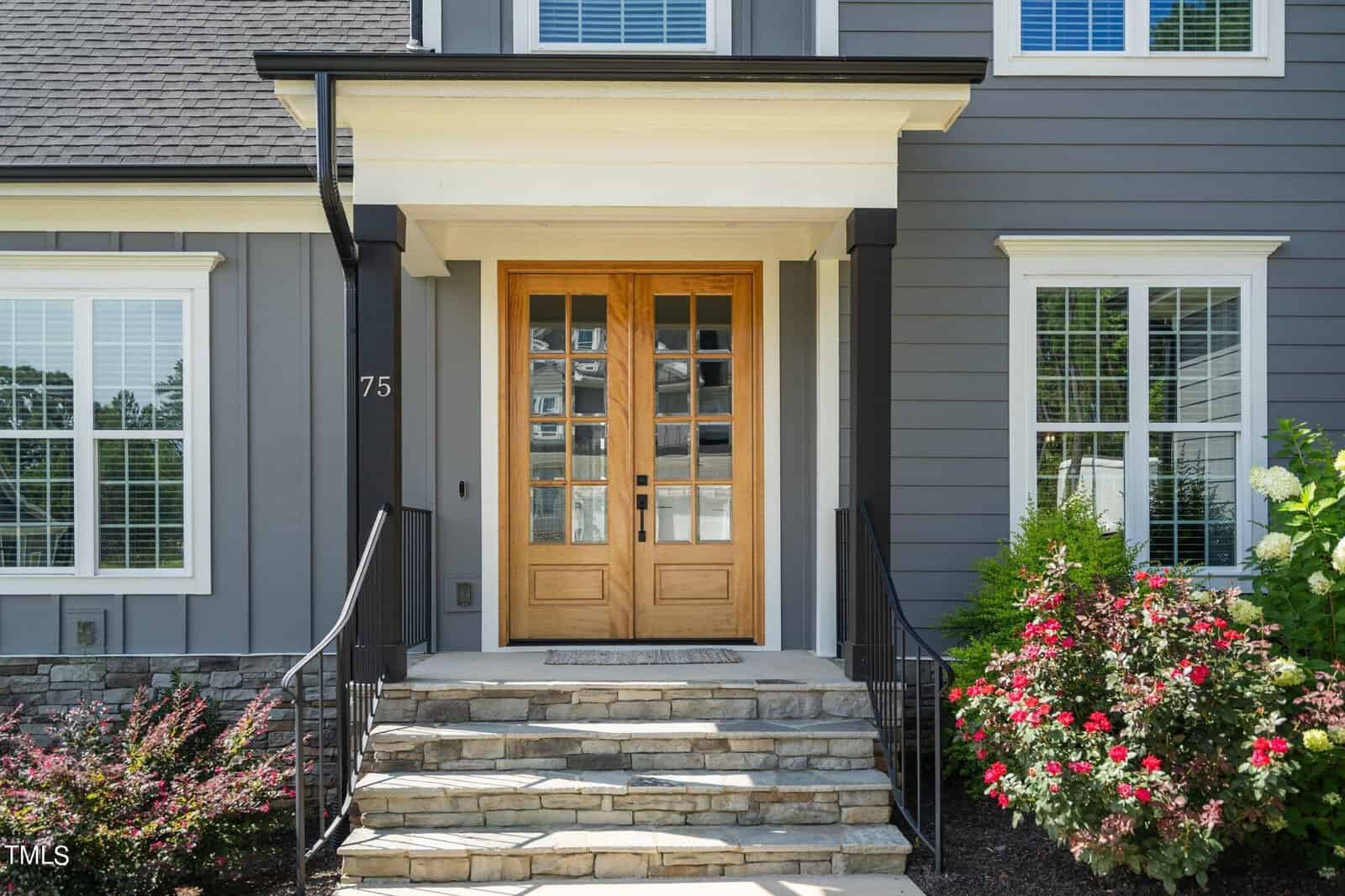 A modern house entrance with double wooden doors, stone steps, black railings, gray siding, and white trim. Colorful flowering bushes and green shrubs are on either side of the steps.