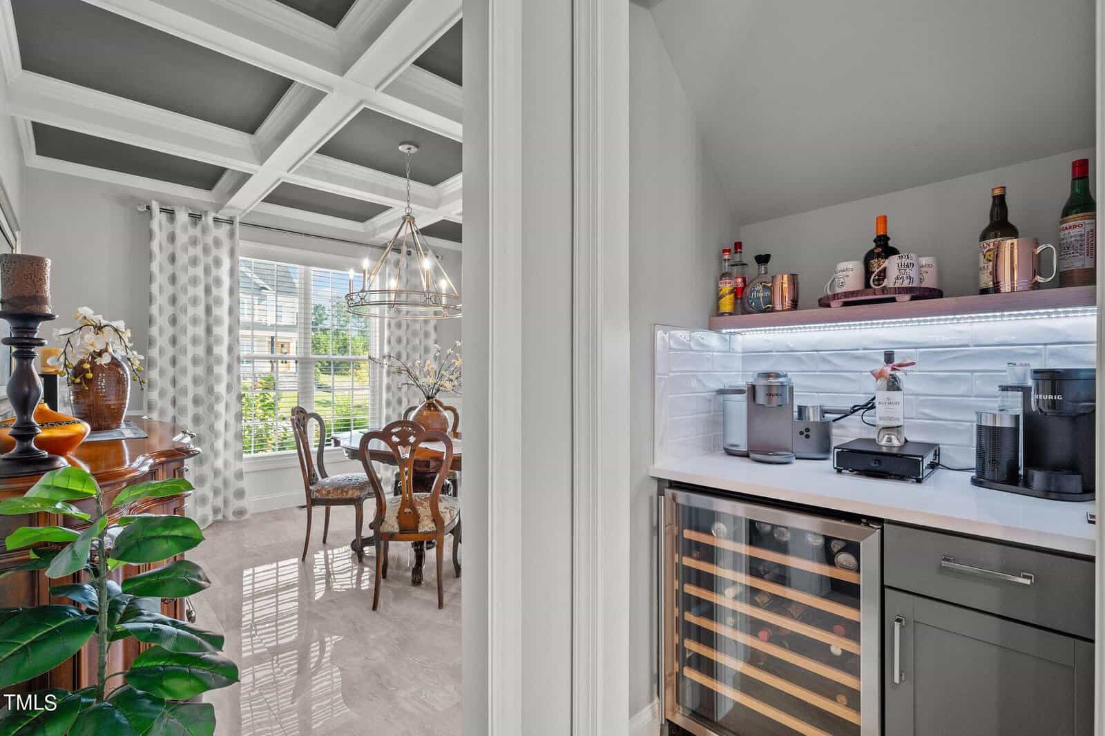 A modern dining room with a wooden table and chairs is visible through a doorway beside a stylish butler's pantry featuring a wine fridge, coffee makers, and liquor bottles on shelves under accent lighting.