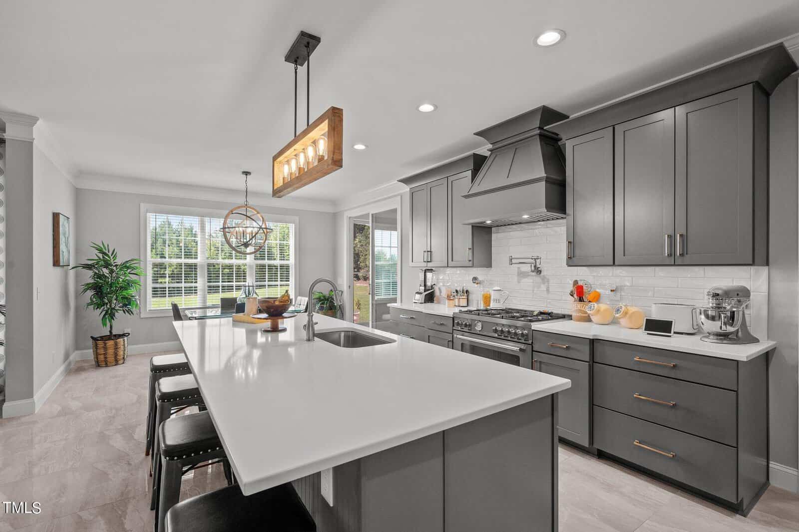 Modern kitchen with gray cabinets, white countertops, stainless steel appliances, large island with sink and stools, pendant lighting, and a window letting in natural light.