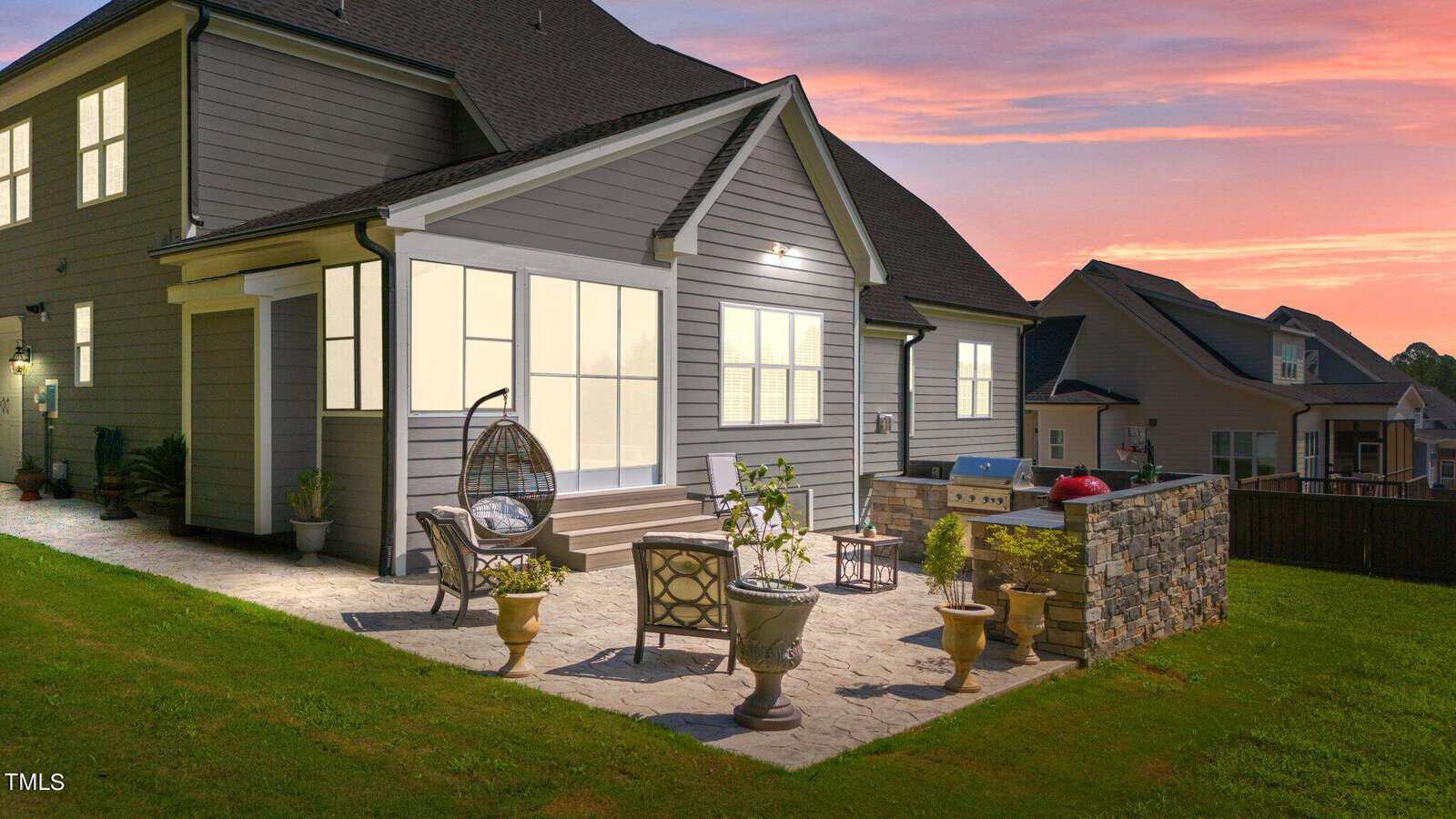 Backyard patio of a modern house at sunset, featuring outdoor furniture, a hanging chair, potted plants, and a stone grill station, with neighboring homes visible in the background.