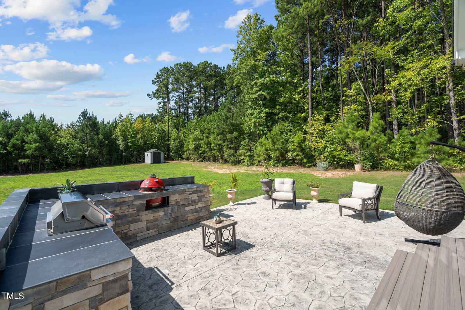 Outdoor patio with a built-in stone grill, red ceramic smoker, cushioned chairs, a hanging egg chair, and a small table. The patio overlooks a spacious grassy yard bordered by dense trees under a blue sky with clouds.