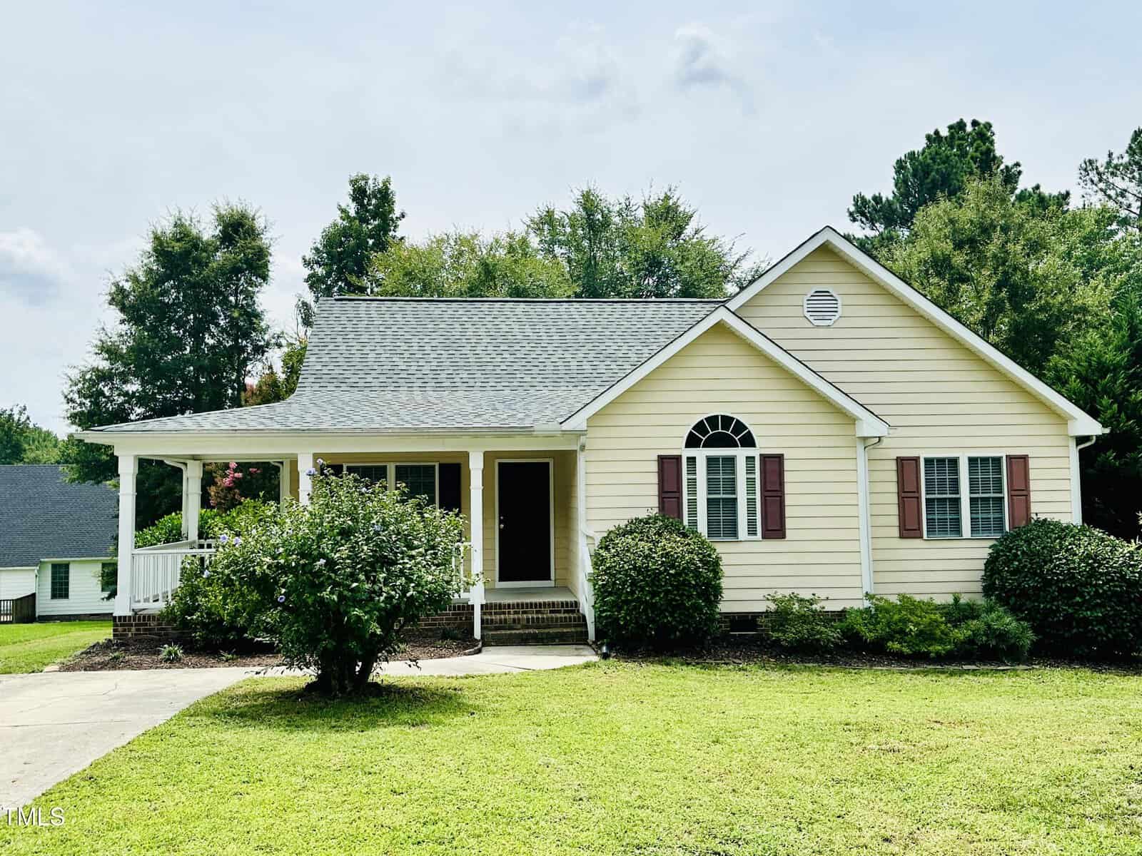 A charming yellow single-story house with white trim stands on 2752 Dorothy Court in Creedmoor. It features a gabled roof, front porch, and red shutters. Surrounded by a green lawn and shrubs, the home is set against a backdrop of trees under a partly cloudy sky.