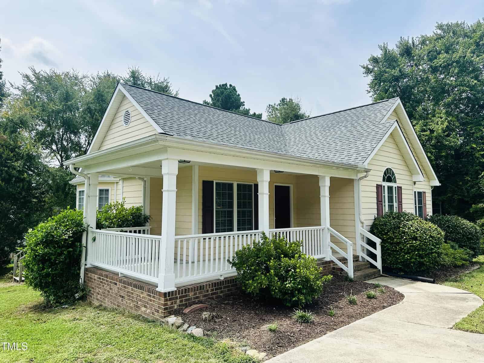 A small, single-story yellow house with a gray shingled roof and a covered porch. Surrounded by green shrubs and trees, it has a concrete walkway leading to the porch entrance. The sky is partly cloudy.