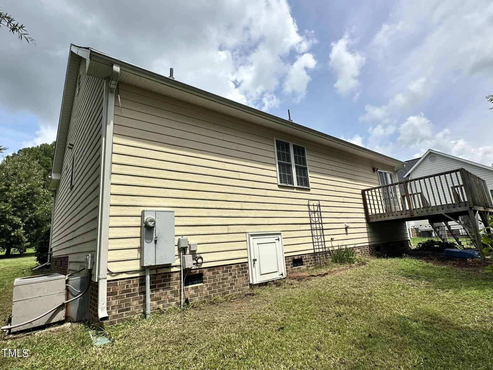 Side view of a single-story house with light-colored siding and a small deck. The house features a window, electrical meter, and an air conditioning unit. The lawn is neatly trimmed and a cloudy sky is visible overhead.