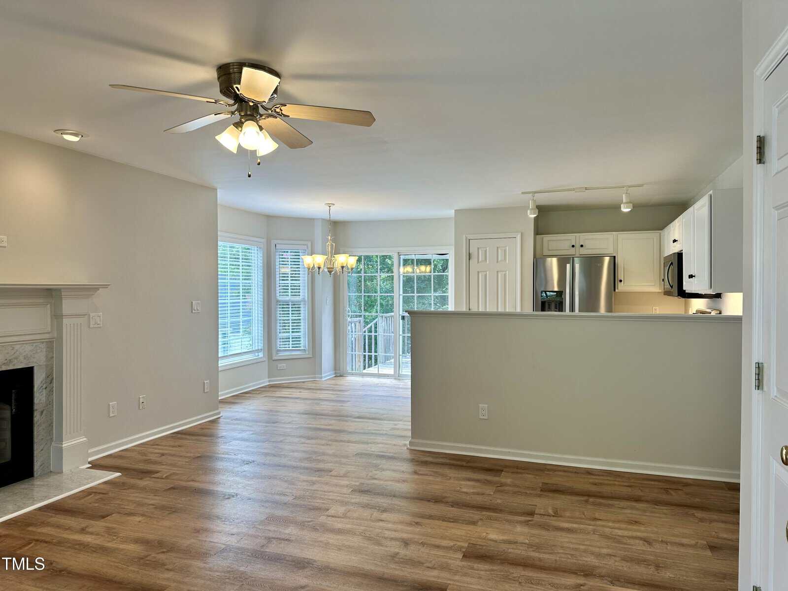 A spacious, open-concept living room with light wood flooring, a ceiling fan, and a fireplace on the left. It leads to a kitchen with white cabinets and stainless steel appliances. Large windows and glass doors provide ample natural light.