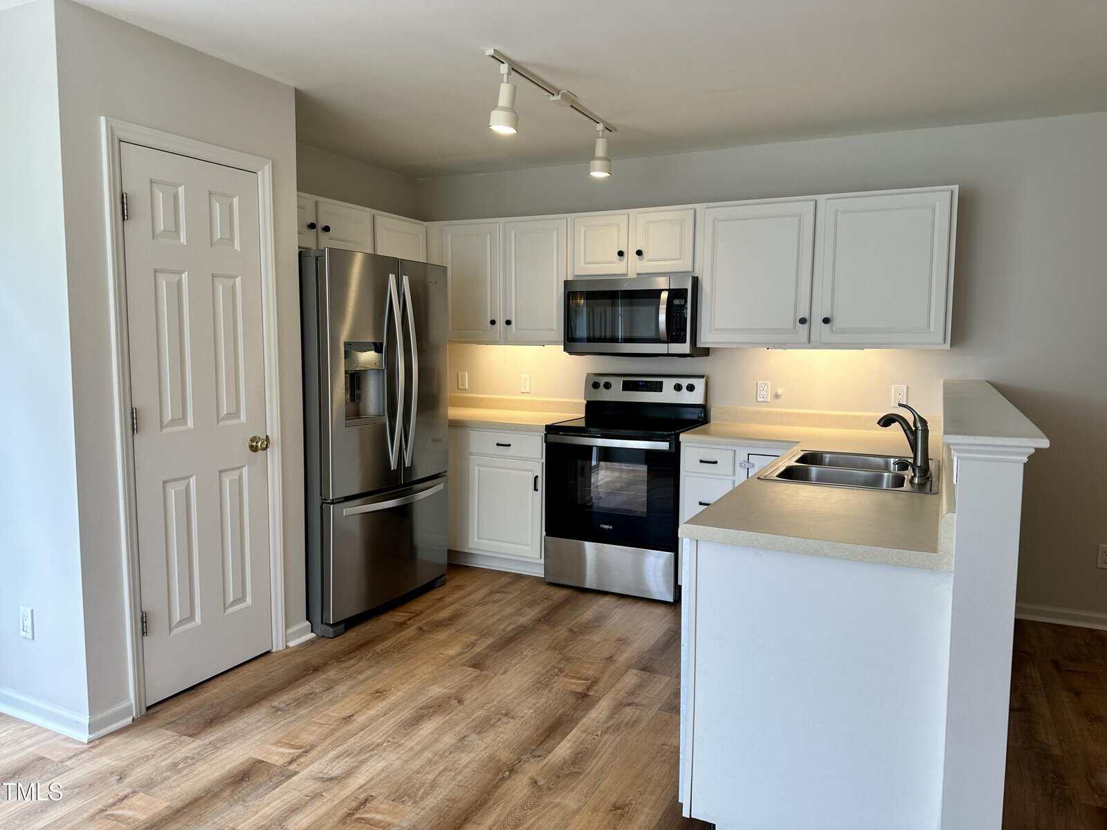 Modern kitchen with white cabinets, stainless steel appliances including a refrigerator, microwave, and oven. Light wood flooring complements the neutral walls. A small peninsula with a sink is on the right, and overhead track lighting is visible.