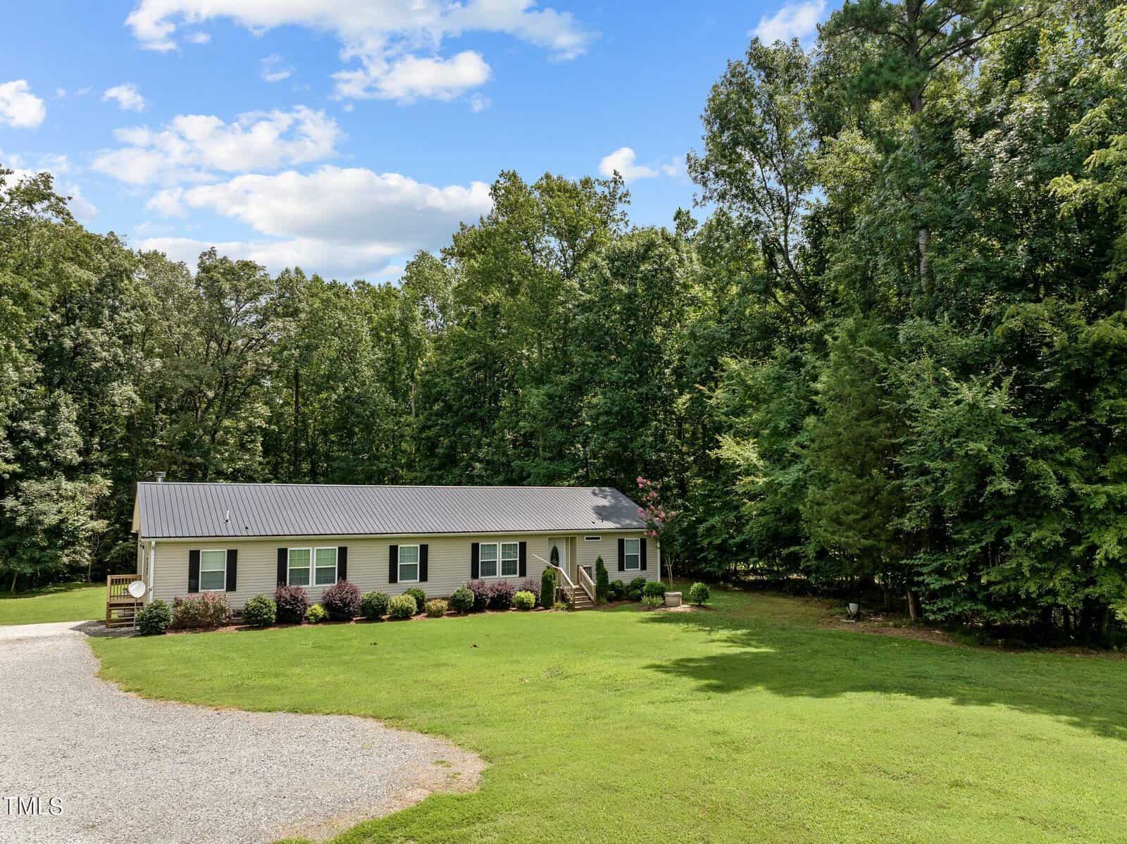 A single-story house with a metal roof surrounded by a lush green lawn and dense trees. The gravel driveway curves toward the front of the house. The sky is bright with a few scattered clouds.