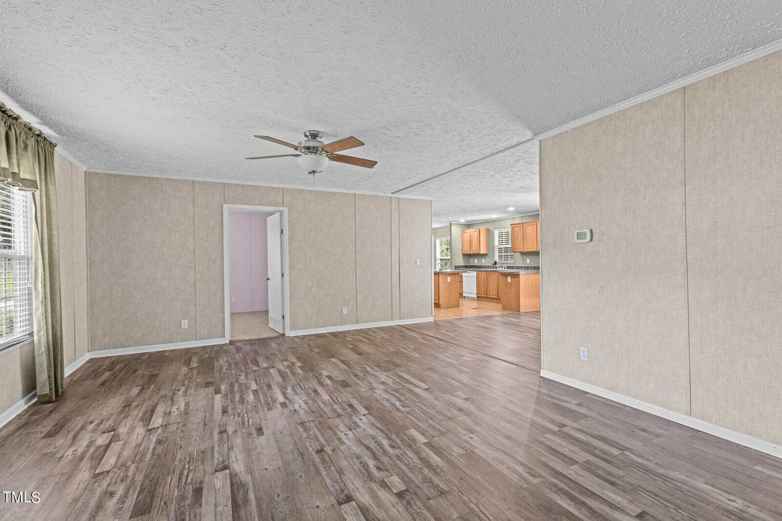 Spacious living room with light-colored walls and wood-look flooring, featuring a ceiling fan. Large windows with green curtains allow natural light. An open doorway leads to the kitchen with wood cabinets in the background.