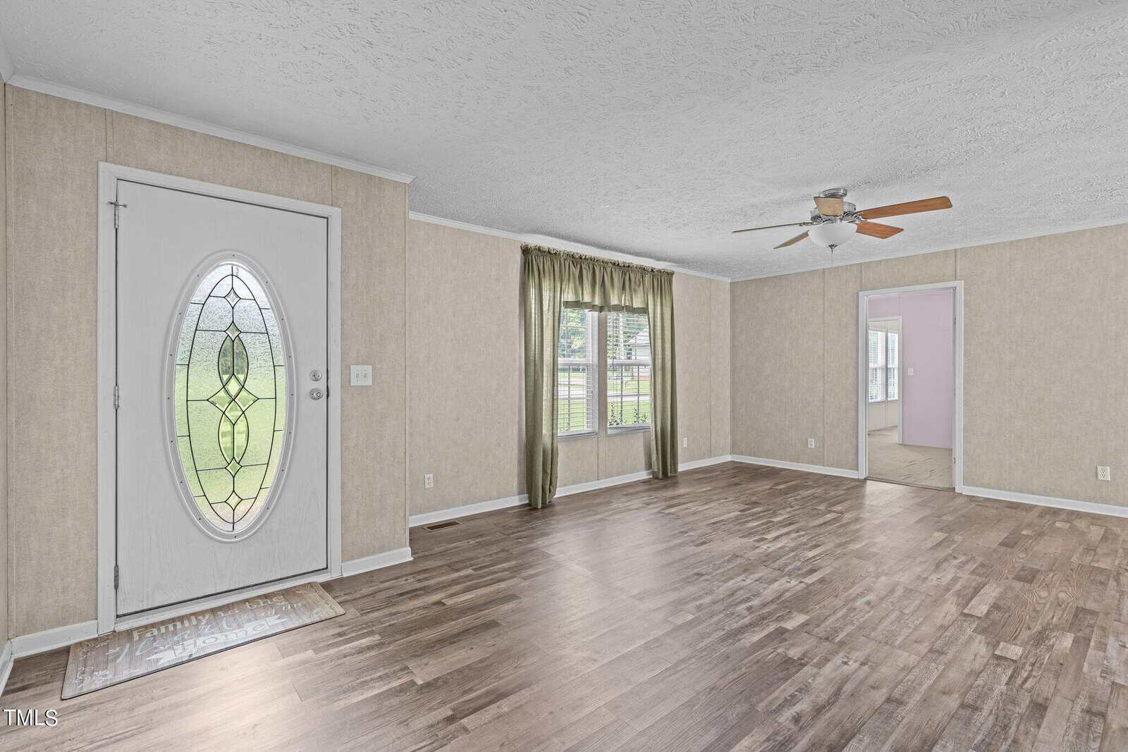 A living room with light wood flooring, beige walls, and a decorative oval glass front door. There's a window with green curtains and a ceiling fan. An open doorway leads to another room.