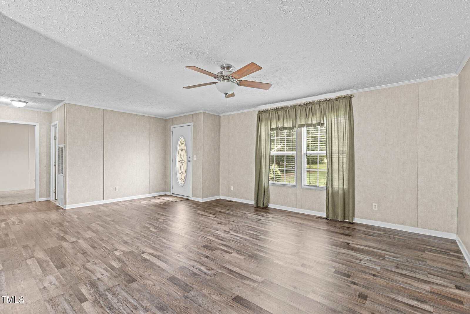 A spacious, empty living room with light-colored walls and wooden flooring. There is a ceiling fan overhead, a window with green curtains on the right, and a decorative oval glass door on the left. Natural light fills the room.