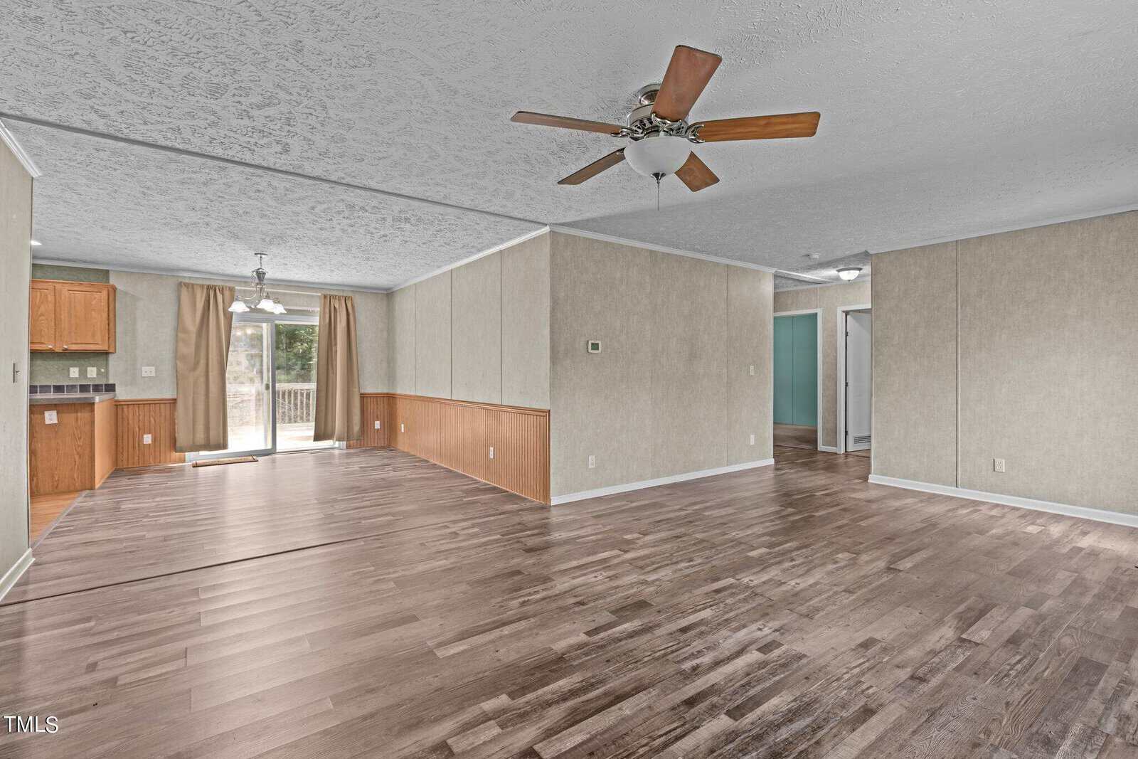 Spacious empty living room with wood flooring, ceiling fan, and recessed lighting. Open layout leads to a kitchen with wooden cabinets and a sliding door opening to a patio. Neutral beige walls complement the space.