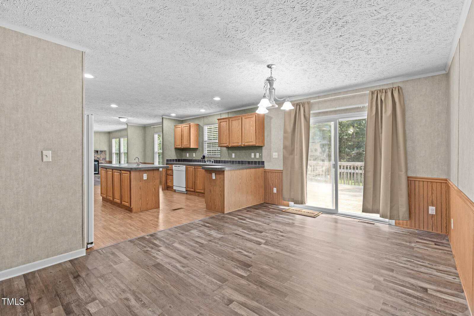 Spacious open kitchen and dining area with wooden cabinets, dark countertops, and hardwood floors. A sliding glass door leads to a deck, allowing natural light to fill the room. Walls have a combination of wallpaper and wooden paneling.