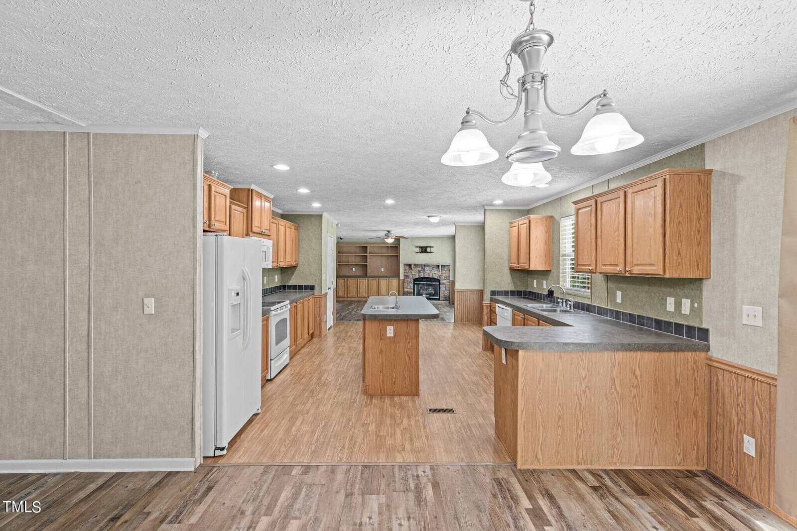 A spacious kitchen with wooden cabinetry and a large island. It features a white refrigerator, stove, and dishwasher. The room is well-lit with a chandelier and recessed lighting, and has a wood floor and light green walls.