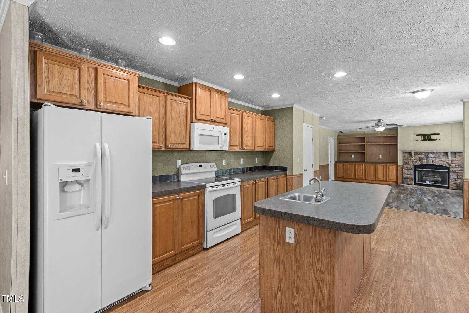 A spacious kitchen with wooden cabinets, a white refrigerator, stove, and microwave. An island with a dark countertop is centered in the room. In the background, there is a cozy living area with a fireplace and a ceiling fan.