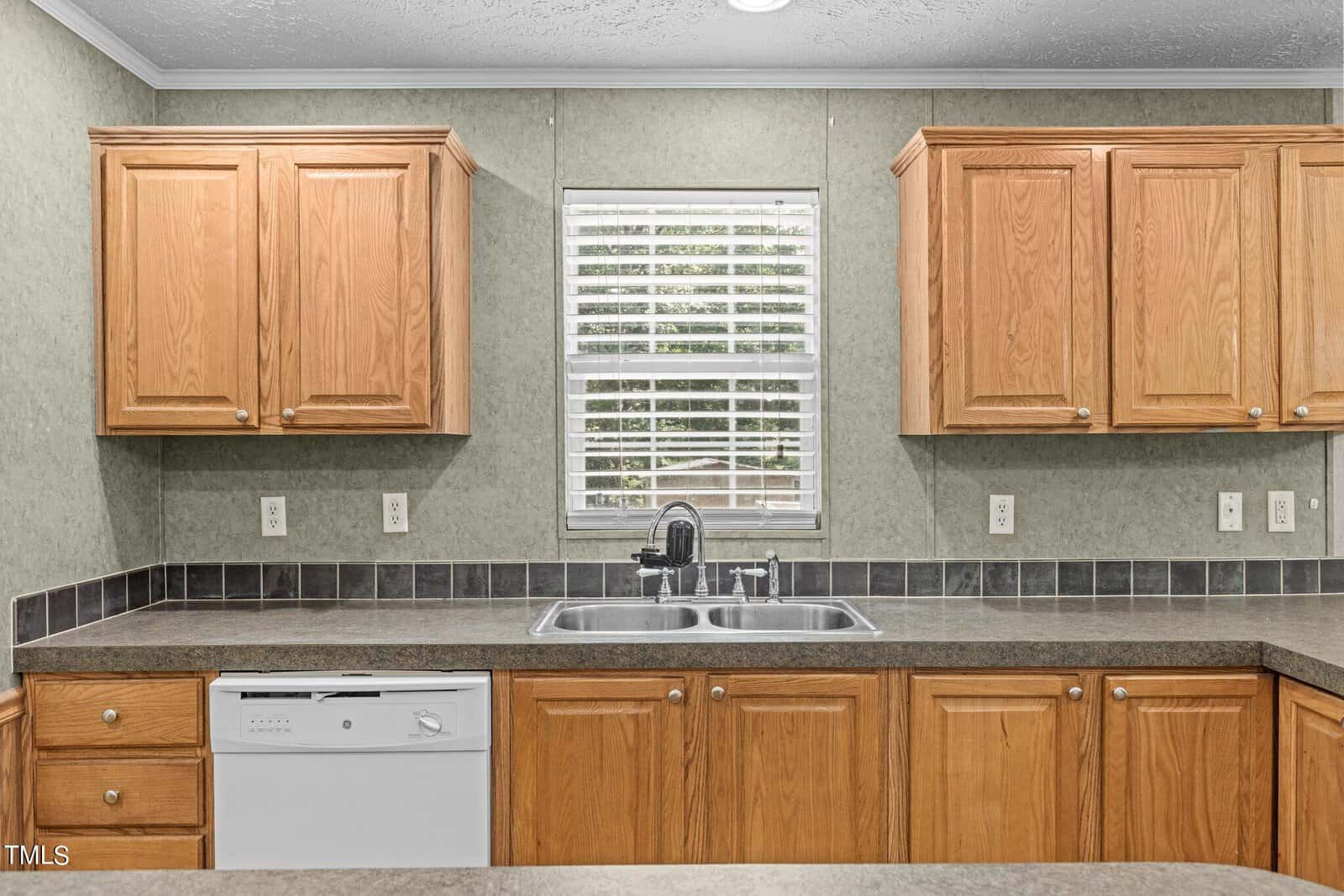 A kitchen with wooden cabinets, a double stainless steel sink under a window with blinds, and a dishwasher. The backsplash is tiled in dark gray with light-colored countertops. The walls are a muted green.