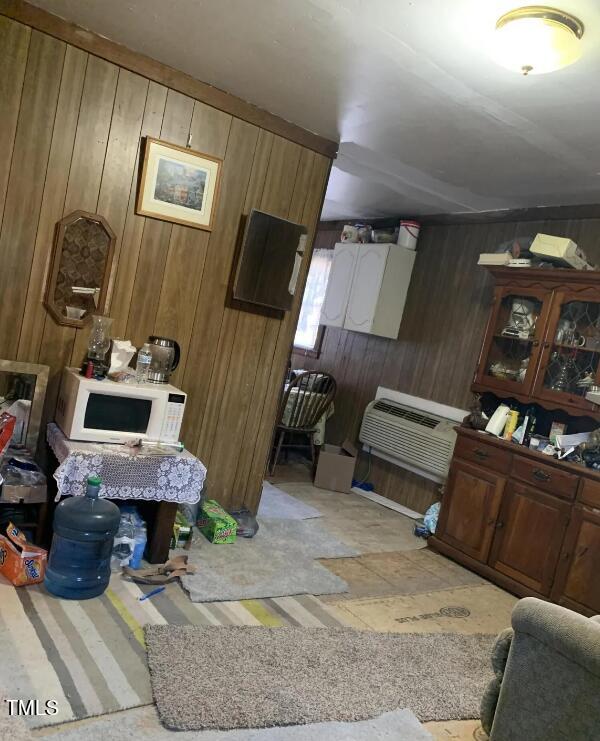 A cluttered room with wood-paneled walls, a microwave on a lace-covered table, assorted items on the floor, a water cooler, and a glass-fronted cabinet filled with items. There is a doorway leading to a small dining area, and an air conditioning unit under the window.