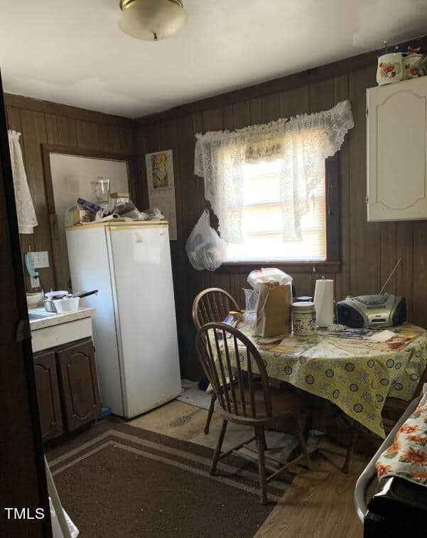 A small, cluttered kitchen with wood-paneled walls, featuring a refrigerator, a table covered with a yellow floral tablecloth, two wooden chairs, and a window with lace curtains. Various items are scattered on the table, and a stereo sits on the counter.