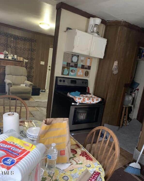 A small, cluttered kitchen and living area. The kitchen has a stove with a pan covered by a cloth, a wall clock above, a table with paper towels, bottled water, fast food bags, foam cups, and miscellaneous items. The living room has a beige recliner and various furniture.