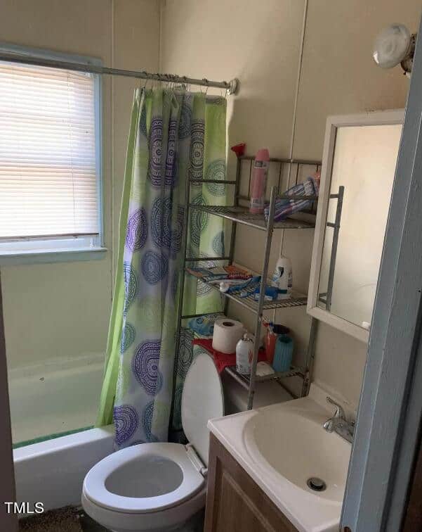 A small bathroom with a white bathtub and shower combination on the left, featuring a green and purple patterned shower curtain. Next to the bathtub is a white toilet with a silver shelving unit above it, stocked with toiletries. A sink with a mirrored cabinet is on the right.