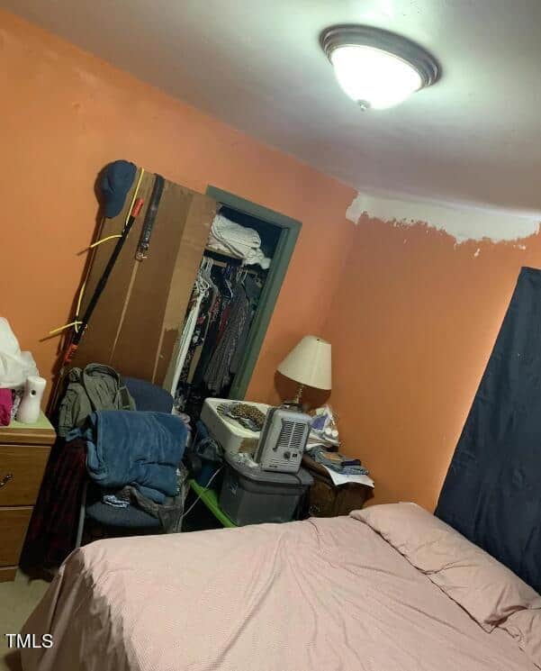 A small bedroom with orange walls, some of which have patches of white. A bed with pink sheets and pillows is positioned next to a cluttered nightstand with a lamp and various items. An open closet shows hanging clothes, and a chair nearby holds more clothing.