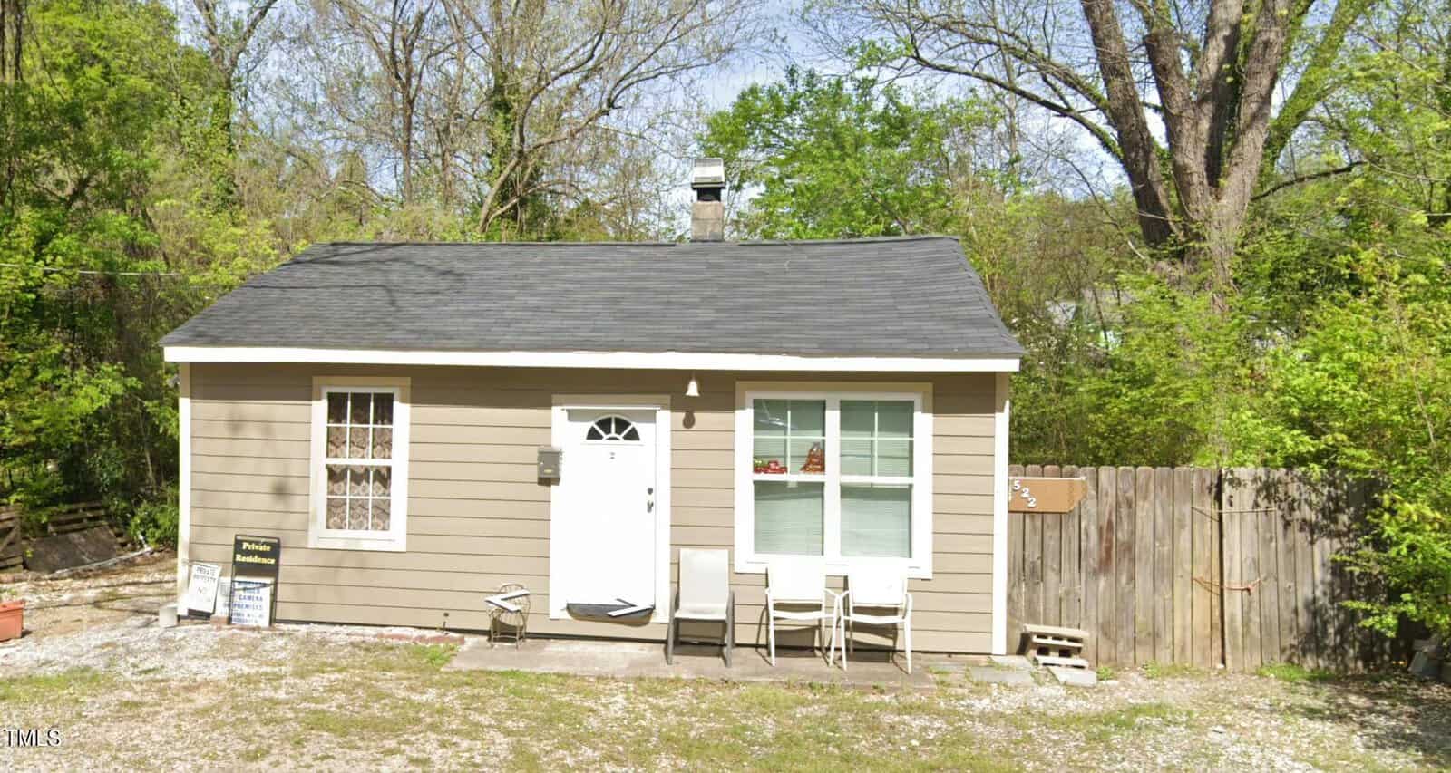 A small, single-story beige house at 522 Hargrove Street in Henderson features a dark gray roof and white trim. It has a white door, two large windows, and a wooden fence to the right. The modest front yard, shaded by trees and brush, hosts two patio chairs for a cozy outdoor retreat.