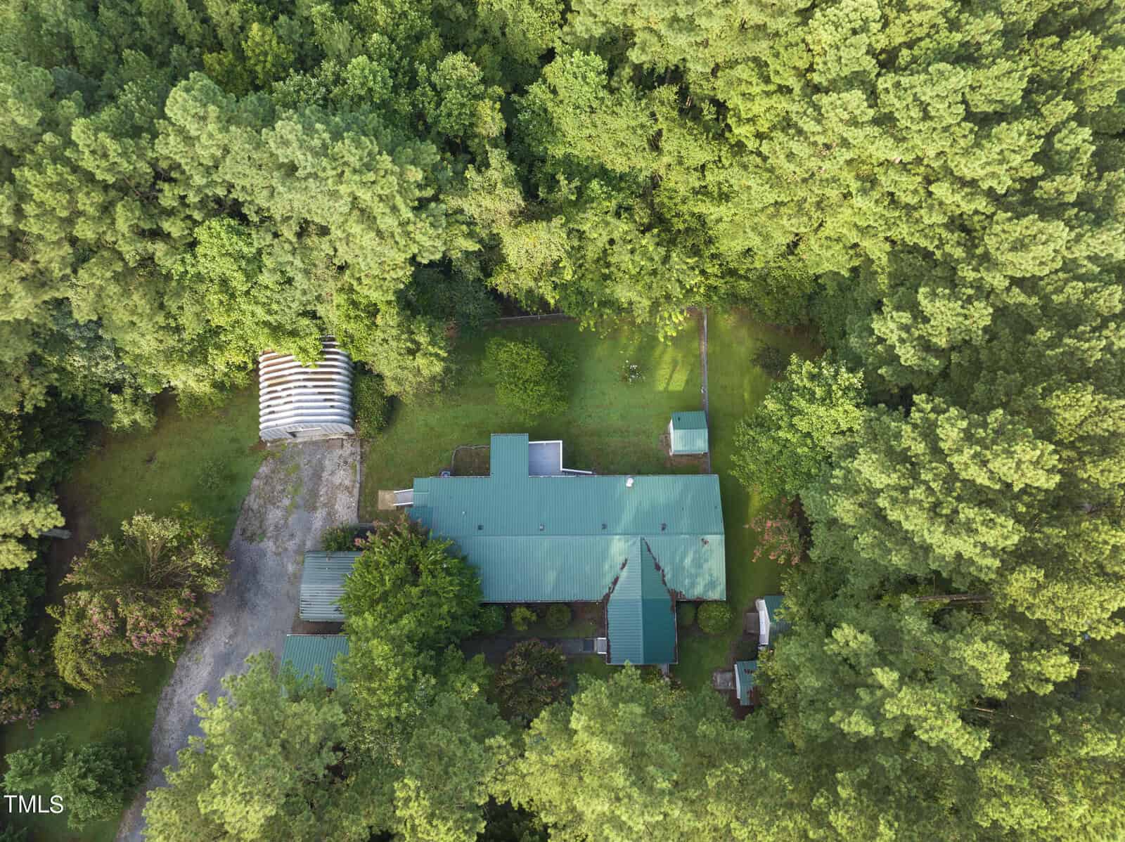 Aerial view of 29 Skylark Lane nestled in a lush, green forest. The main structure has a green roof and is surrounded by tall trees. A smaller building and a covered parking area are to the left, while a grass lawn and driveway are also visible in this Henderson property.