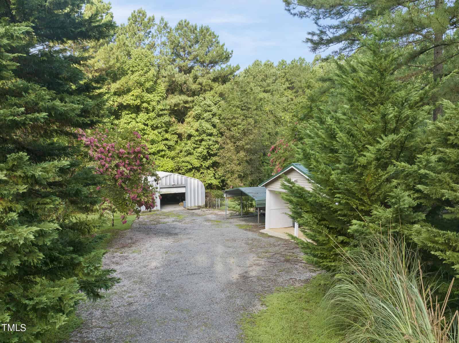A gravel driveway flanked by lush green trees and bushes leads to a covered carport structure and a small building with a green roof. In the background, more trees and a mostly clear sky are visible. The area appears serene and shaded.