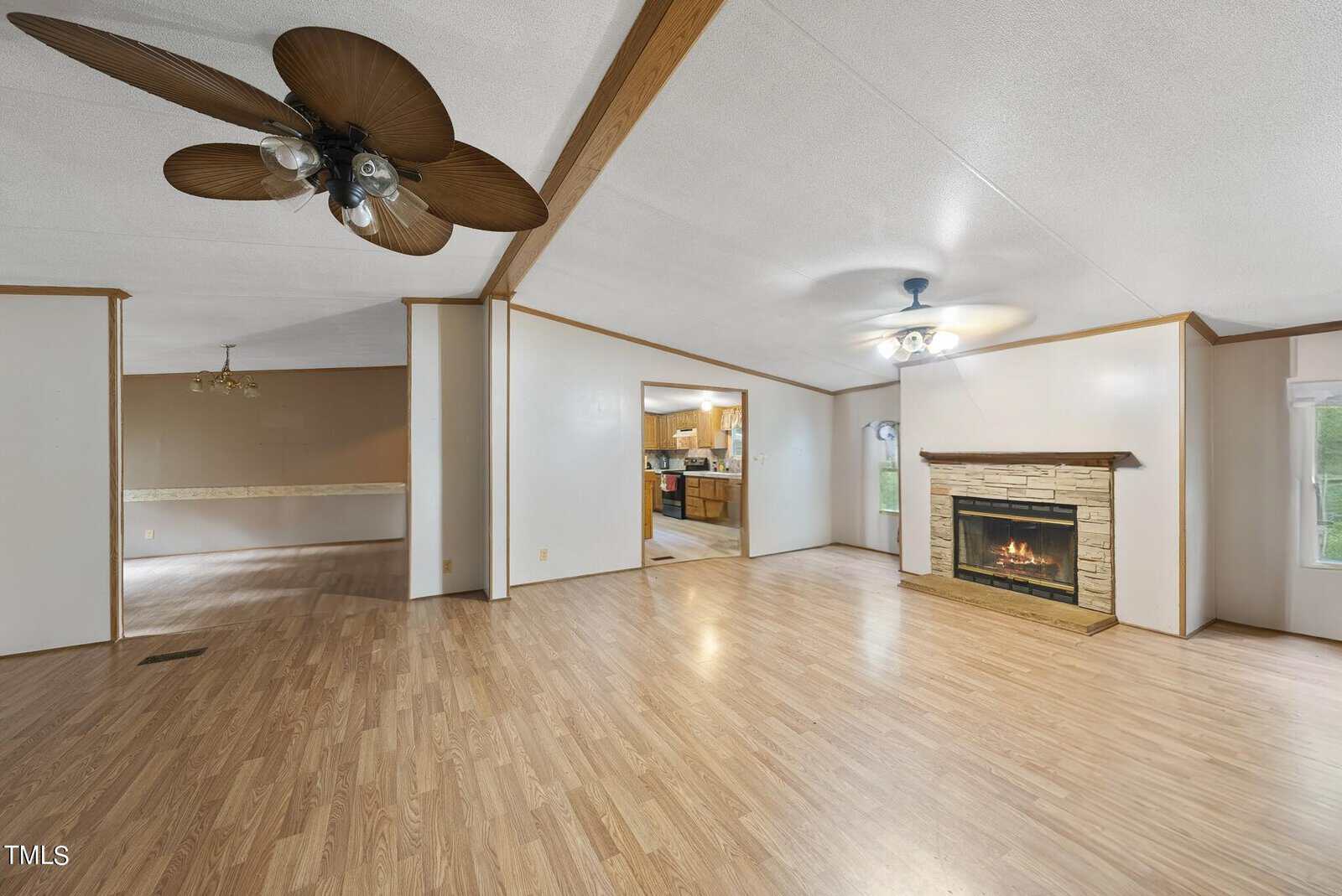 A spacious living area with light wood laminate flooring, a ceiling fan with wooden blades, and a stone fireplace with a fire burning. The room opens into a kitchen with visible appliances and a dining area in the background. Large windows allow natural light to fill the space.