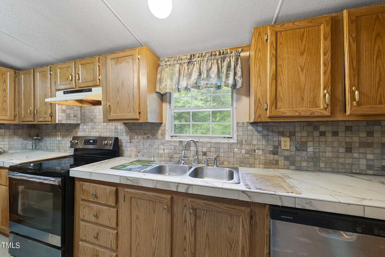 A kitchen featuring wooden cabinets, a double-bowl stainless steel sink beneath a window with floral curtains, an electric stove with an oven, a dishwasher, and a marble-patterned countertop. The backsplash has a mosaic tile design in neutral colors.