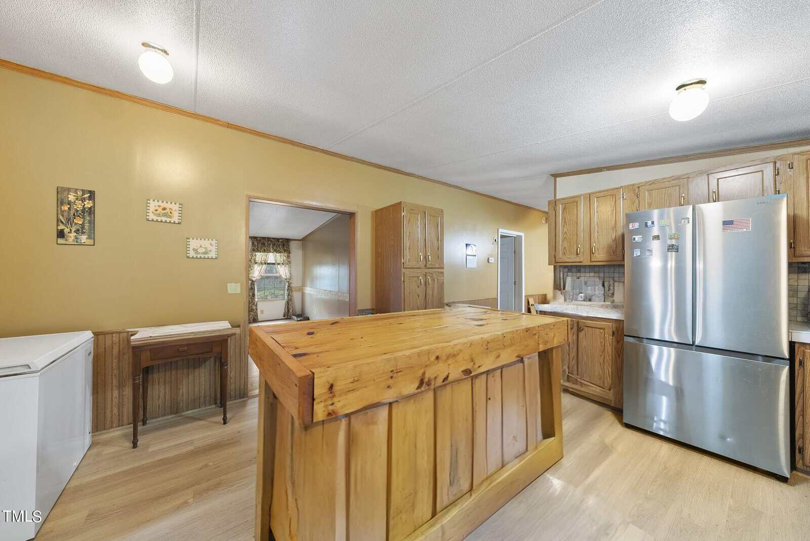 A bright kitchen with wooden cabinetry and a large wooden island in the center. Stainless steel refrigerator stands to the right. Light wood flooring complements the light brown walls. Various wall decor items and a doorway leading to another room visible.
