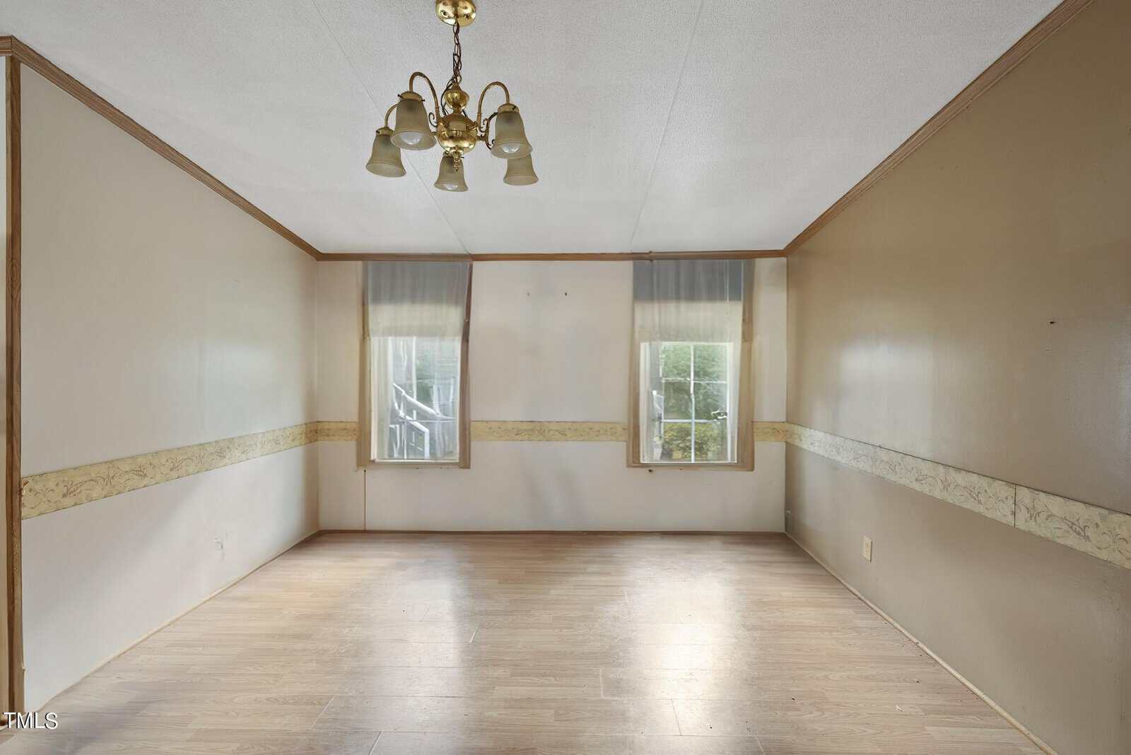 An empty room with beige walls, light wood flooring, two windows with sheer curtains, and a brass chandelier hanging from the ceiling. A strip of decorative wallpaper runs horizontally along the middle of the walls.
