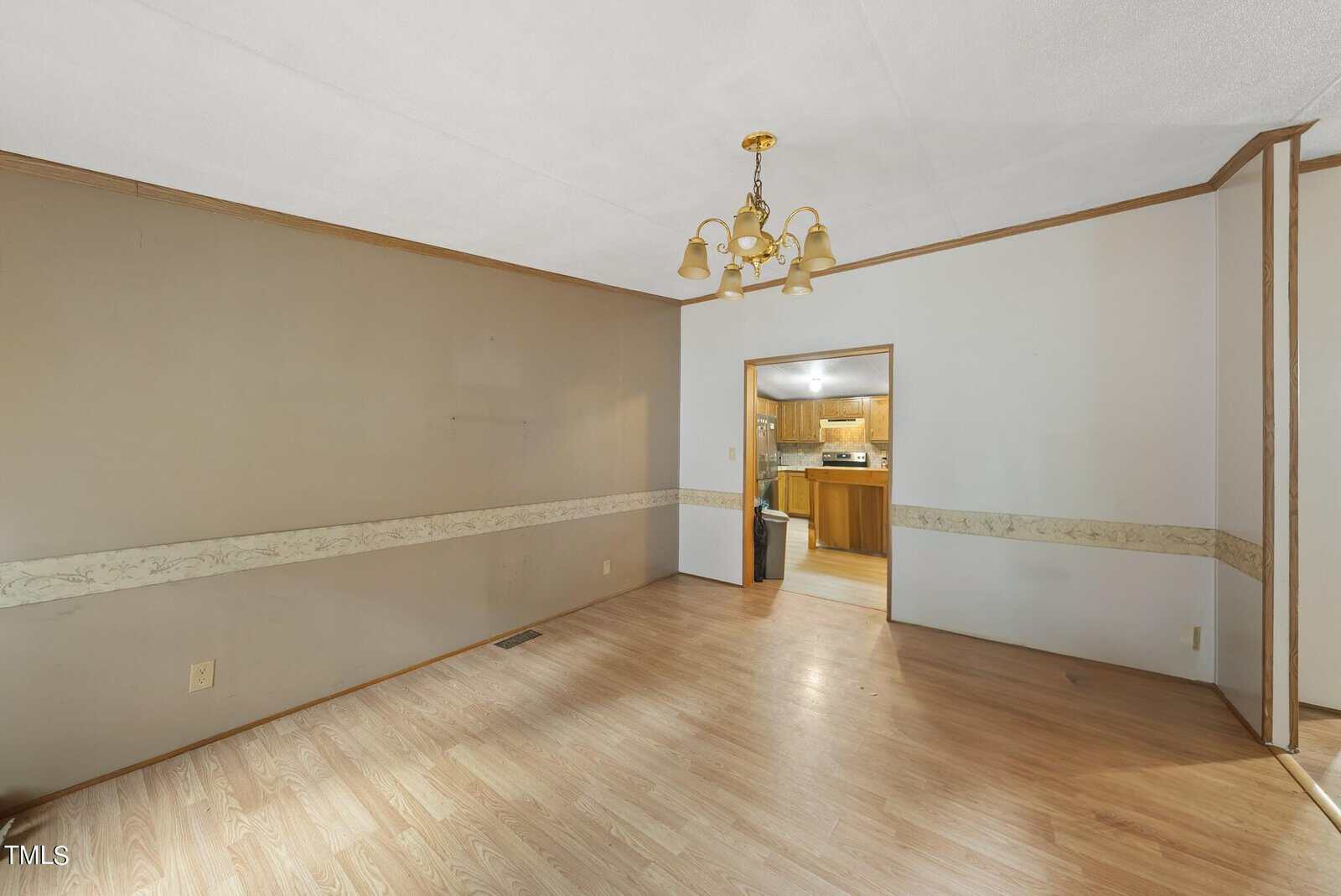 A mostly empty room with light wooden flooring and beige walls featuring a decorative border. The room contains a ceiling-mounted, gold-toned chandelier. There's an open doorway leading to a kitchen area with wooden cabinets and visible appliances in the background.