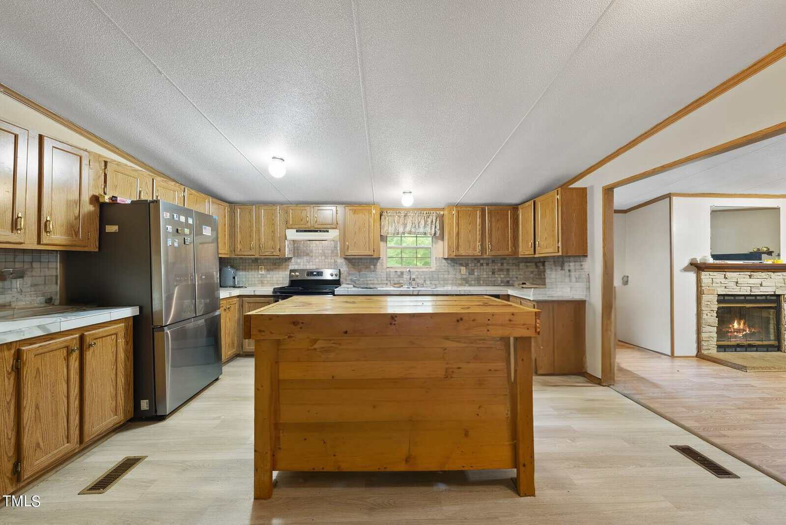 A spacious kitchen with wooden cabinets and a large island in the center. The appliances include a stainless steel refrigerator and oven. The backsplash features a light tile design, and there's a small window above the sink. A cozy living room with a fireplace is adjacent.
