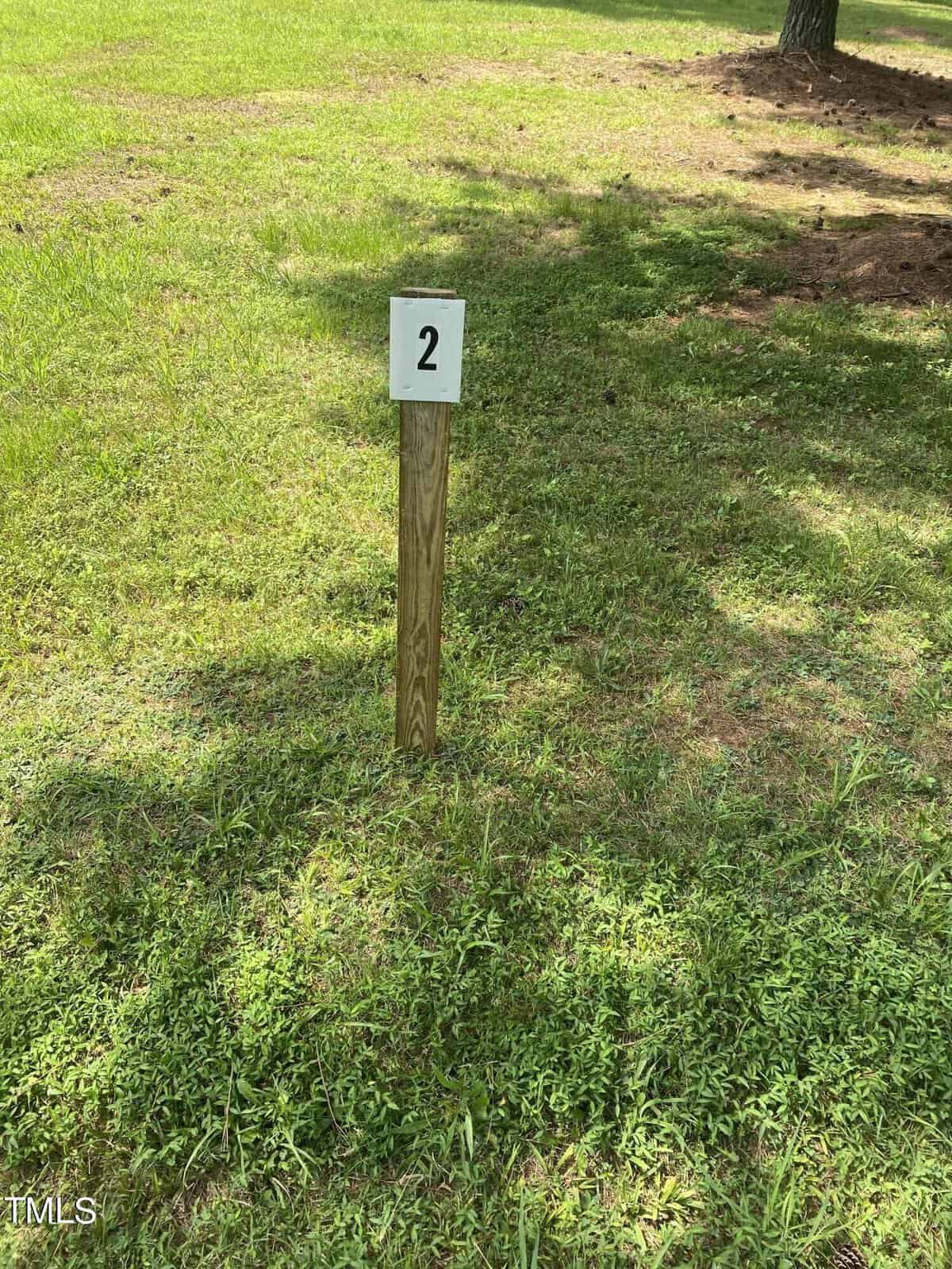 A wooden post with a white square on top displaying the number "2" stands on a grassy field. Sunlight casts shadows on the grass, with sparse patches of earth visible.